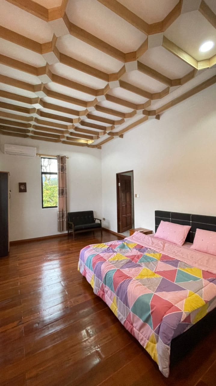 A brightly lit sleeping area features a spacious bed covered with a colorful geometric-patterned blanket. Wooden floors complement the elegant coffered ceiling. A small seating area is visible in the corner, and natural light enters through a large window adorned with sheer curtains.