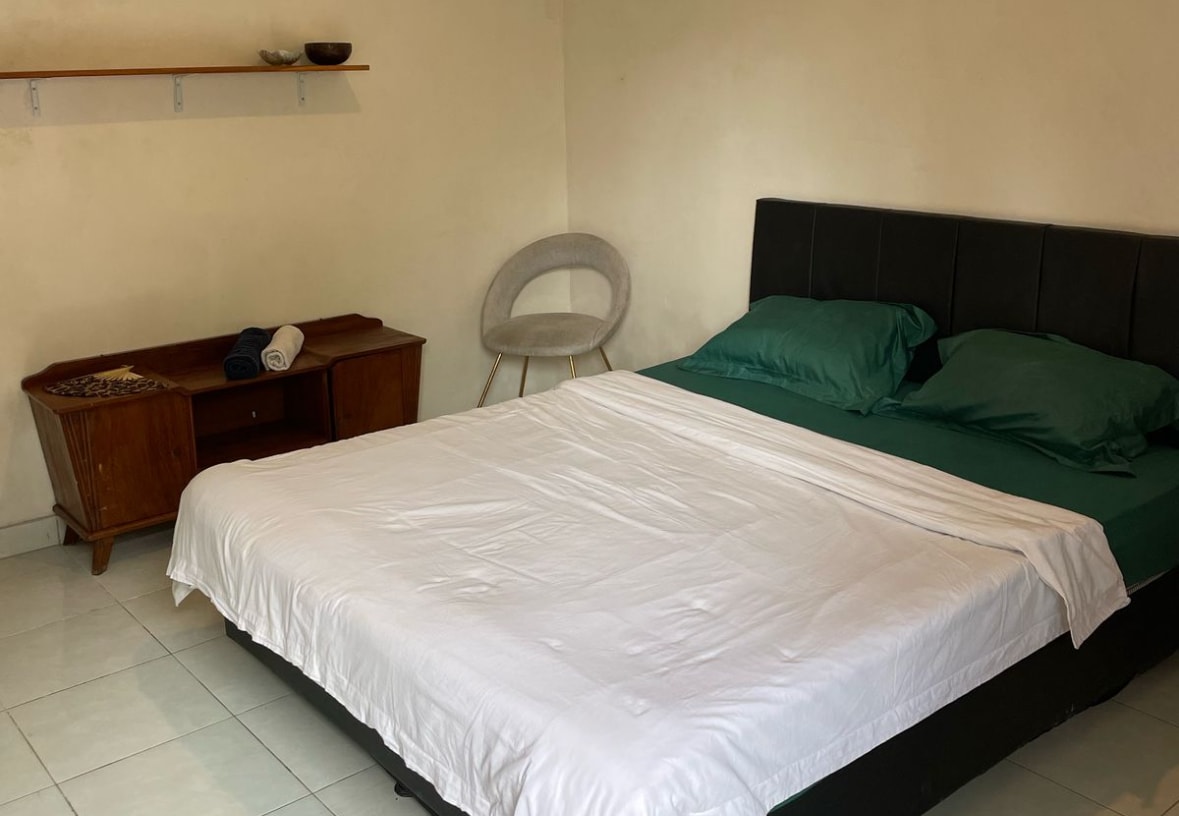 A spacious bed with dark green bedding is positioned in a simple room with white tile flooring. A sleek, light-colored chair is placed to one side, while a wooden dresser stands against the wall, providing a functional space for personal items. Natural light is present.