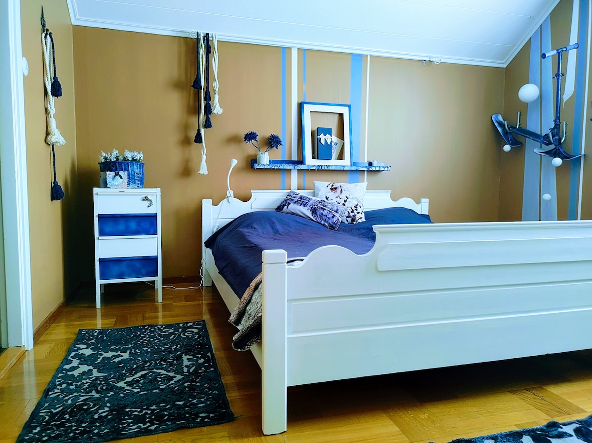 A bedroom features a spacious bed with dark bedding positioned against a light brown wall. A bedside table with a blue accent stands nearby. Decorative elements such as wall art and tassels add character. A patterned rug lays on the polished wooden floor.