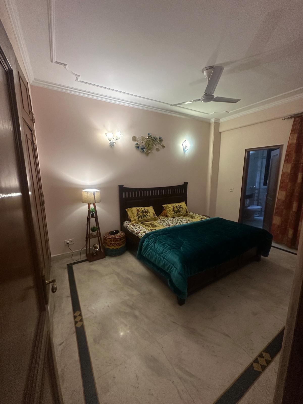 A serene bedroom is featured, showcasing a spacious bed adorned with vibrant green bedding and decorative pillows. A lamp stands beside the bed, adding a soft glow. A mirror and window are visible, and the room is finished with polished marble flooring.