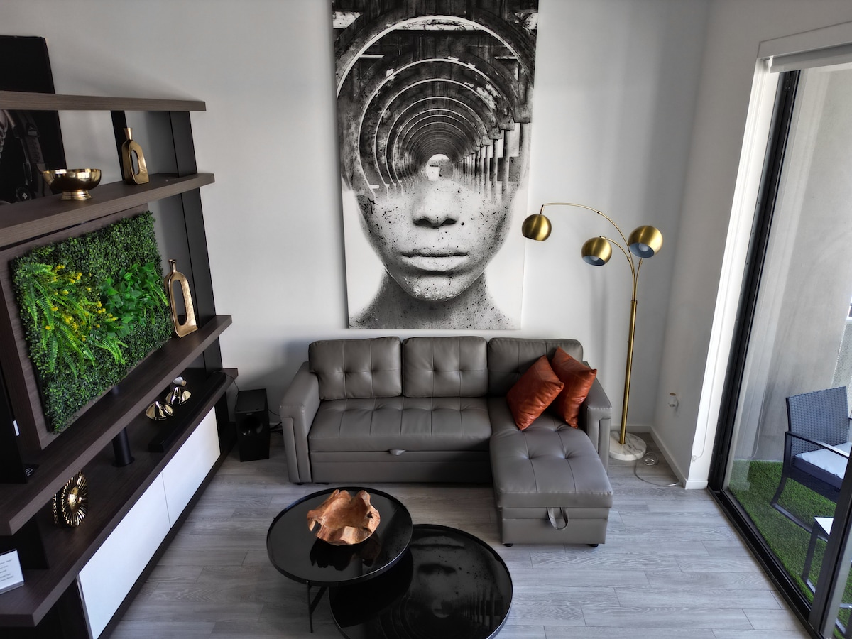 A modern living area features a stylish gray sofa with an orange accent pillow, positioned next to a sleek black coffee table. A television is mounted on a decorative wall shelf that includes greenery, while a floor lamp adds light. Large glass doors open to the balcony.