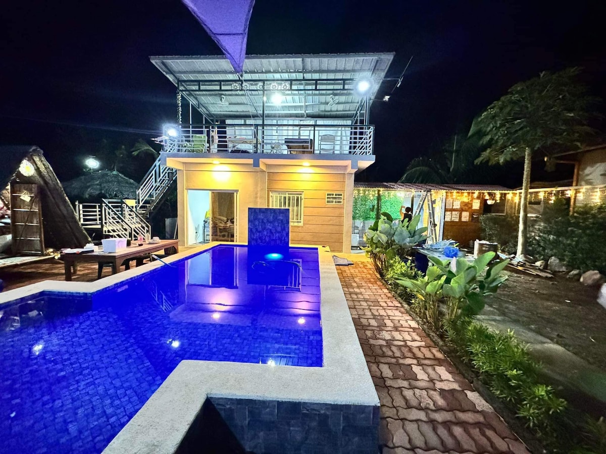 A modern two-story villa is highlighted at night, featuring a shimmering blue pool with ambient lighting. Lush greenery surrounds the outdoor space, and pathways are lined with decorative stones. The upper level balcony overlooks the pool area, offering an inviting social space.
