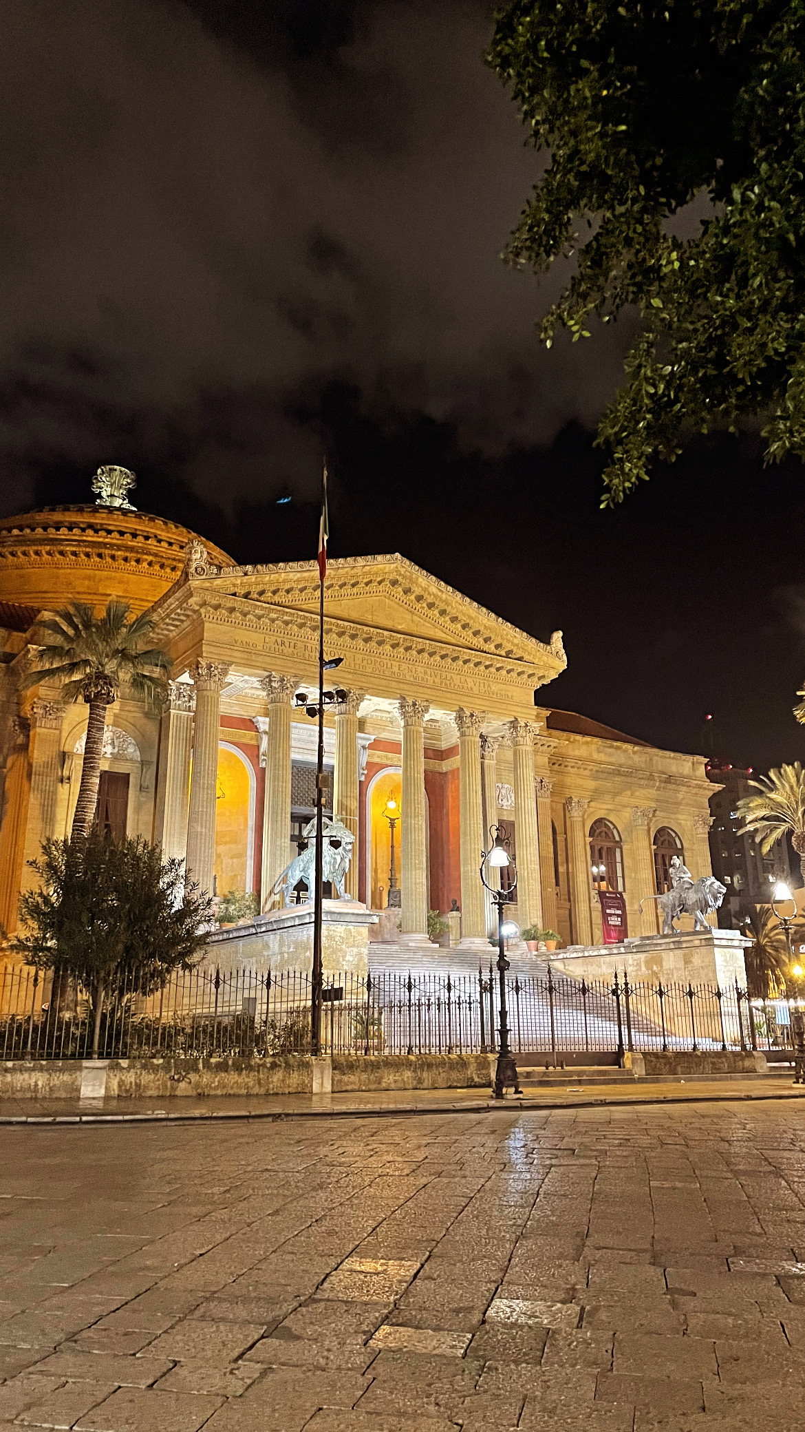 The Teatro Massimo, an architectural landmark, is illuminated at night, showcasing its grand entrance with steps flanked by statues. Tall palm trees are visible nearby, while the vibrant façade reflects warm tones under the night sky, contributing to the lively atmosphere of Palermo.