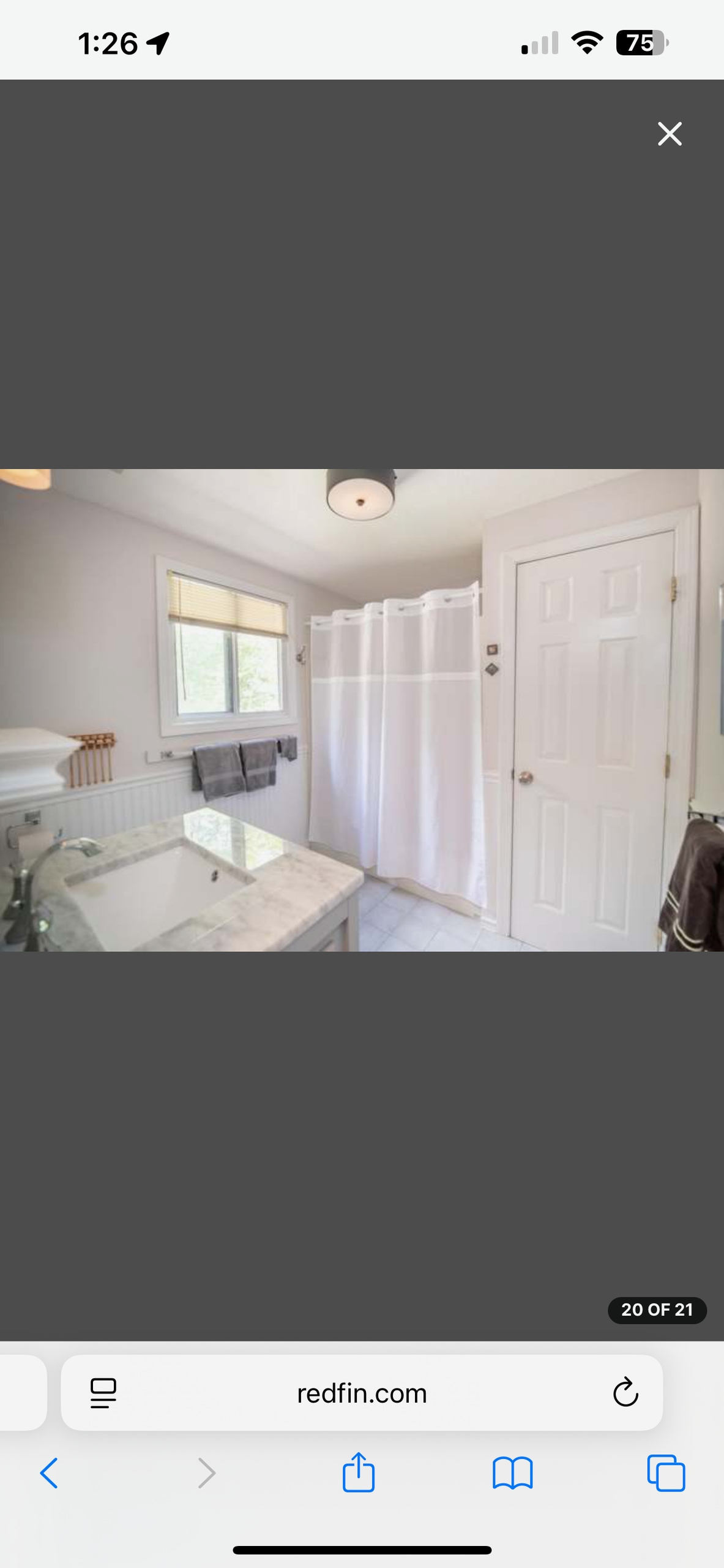 A full bathroom is presented with a marble countertop and a spacious tub. A white shower curtain separates the bathing area from the rest of the room. Natural light enters through a window above the sink, enhancing the clean, bright atmosphere.