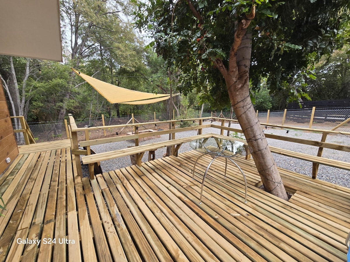 A wooden terrace features spacious seating arrangements and a glass table, surrounded by greenery. A canopy is draped overhead, providing shade, while a nearby tree enhances the natural setting. The area is designed for relaxation and enjoyment of the outdoor environment.