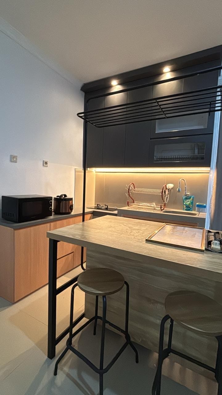 A modern kitchen area features sleek cabinetry and illuminated countertops. A microwave and small appliances are visible on a wooden base, complemented by a spacious island with two stools. Organized kitchenware is neatly arranged, adding to the functional design.