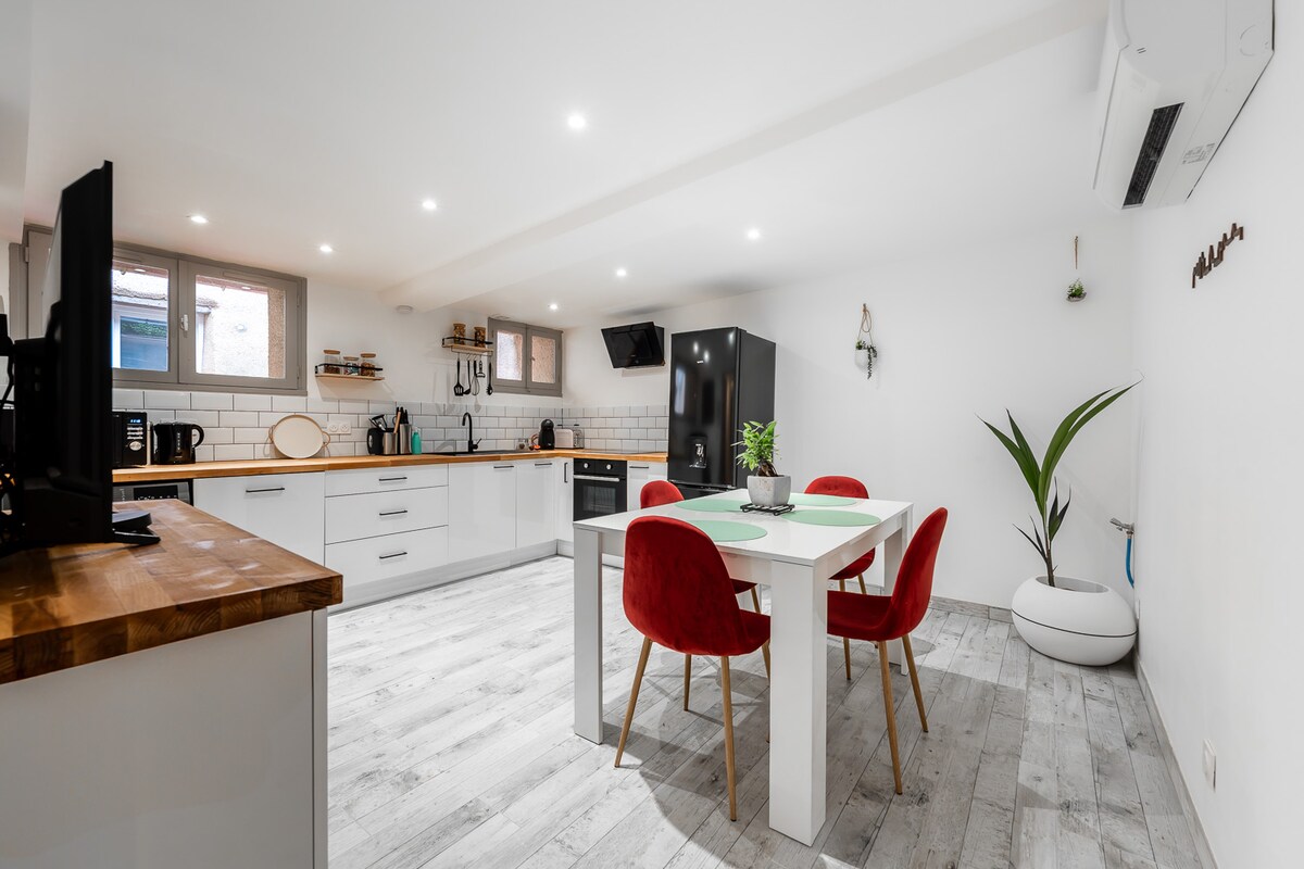 A well-equipped kitchen and dining area features a central white dining table surrounded by red chairs. Modern cabinets and appliances are visible, while light wood accents and neutral walls create a bright, inviting space. Air conditioning and a potted plant add a touch of comfort.