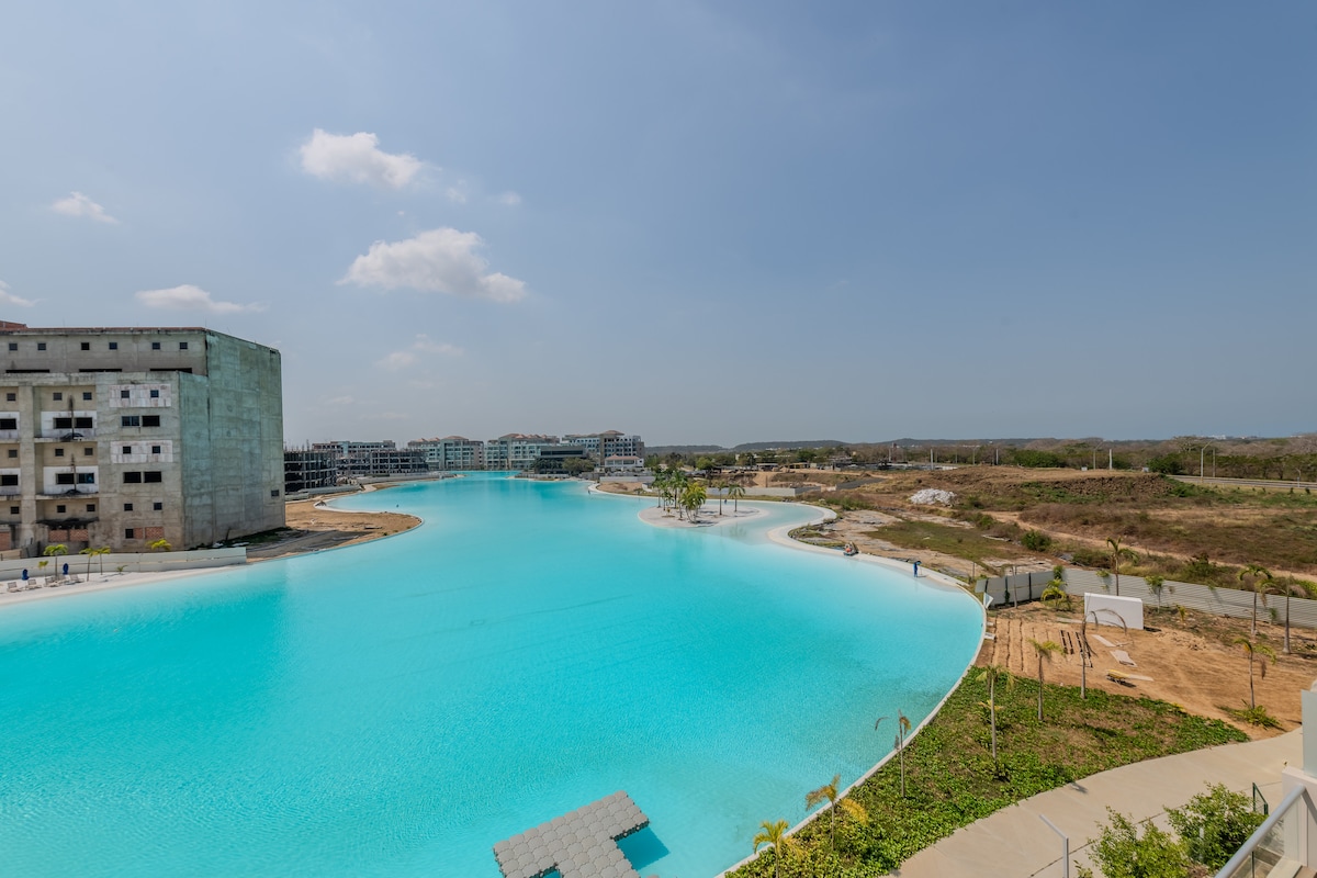 A serene view of the Crystal Lagoon is showcased, featuring vibrant turquoise waters under a clear sky. The surrounding area includes partially constructed buildings, greenery, and a pathway. Sunlight reflects off the water, enhancing the tranquil atmosphere of this inviting environment.