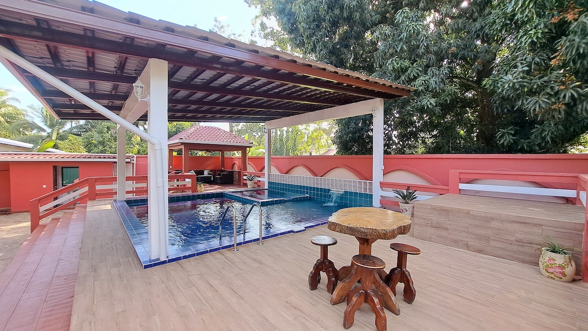 An outdoor area features a swimming pool surrounded by a wooden deck. A shaded structure offers seating and a dining space, complemented by tropical greenery and colorful walls. Stone tables and wooden stools provide an inviting setting for relaxation and gatherings.