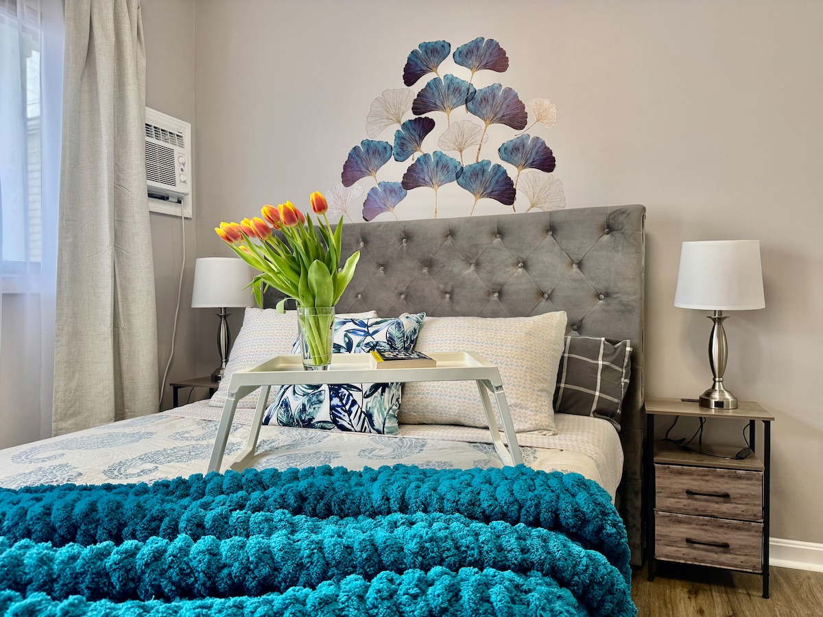 A cozy sleeping area features a tufted gray headboard with decorative pillows arranged on a spacious bed. A vibrant bouquet of tulips rests on a breakfast tray, and a soft teal throw blanket adds warmth. Soft natural light filters through sheer curtains, complementing the serene decor.