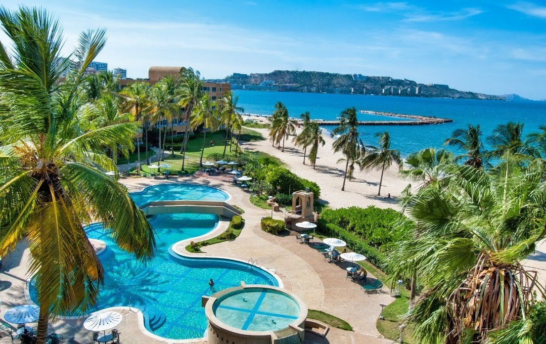 The image features a serene outdoor space, showcasing a swimming pool surrounded by palm trees and lounge chairs. A sandy beach extends into the ocean, with gentle waves lapping at the shore. Clear blue skies enhance the tranquil atmosphere of the scene.