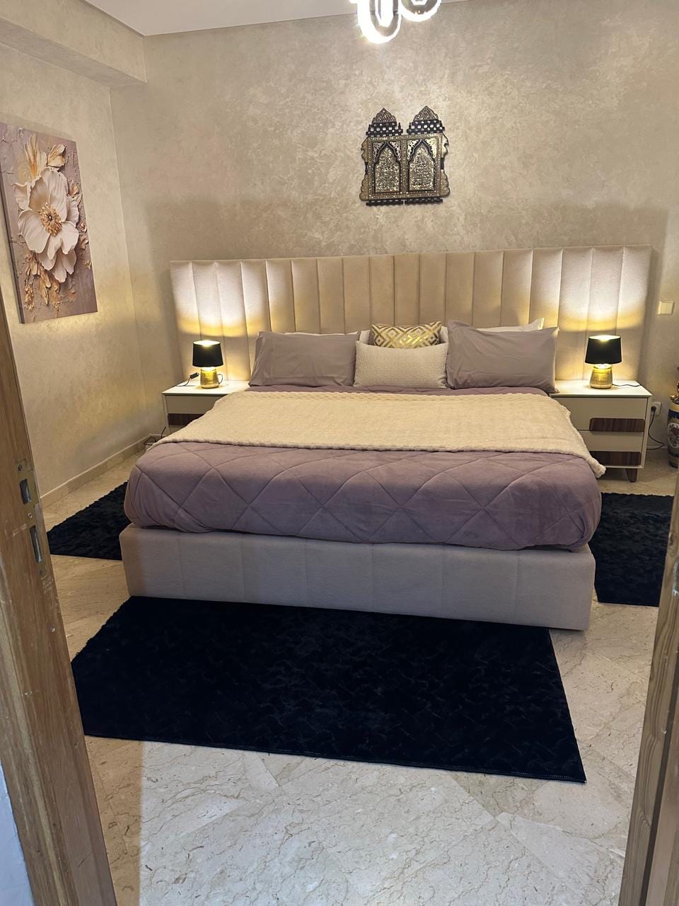 A spacious bedroom features soft taupe walls and elegant decorative elements. A plush bed with a light-colored quilt is positioned centrally, flanked by two bedside tables with lamps. Dark rugs provide contrast against the marble floor, enhancing the inviting atmosphere.