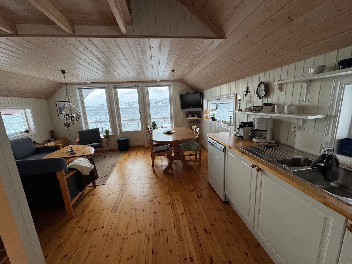 The open living area features large windows allowing natural light to fill the space, offering views of the water. A dining table is set for four, while a comfortable sofa and small side table create a cozy seating area. Wooden cabinetry and countertops are visible in the kitchen area.