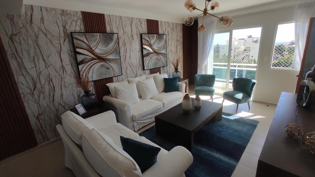 A modern living space features a white sofa with coordinating cushions, surrounded by two teal armchairs. A dark wood coffee table sits on a textured area rug. Light enters through large windows, accentuating the marble wall art and warm décor.