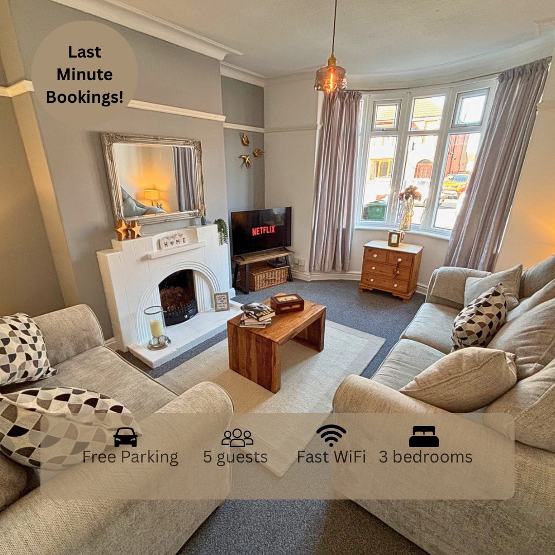 The living area features a cozy seating arrangement with two light grey sofas and a wooden coffee table. A vintage-style fireplace is positioned beneath a decorative mirror. A television with streaming capabilities is visible, and natural light enters through large windows adorned with light curtains.