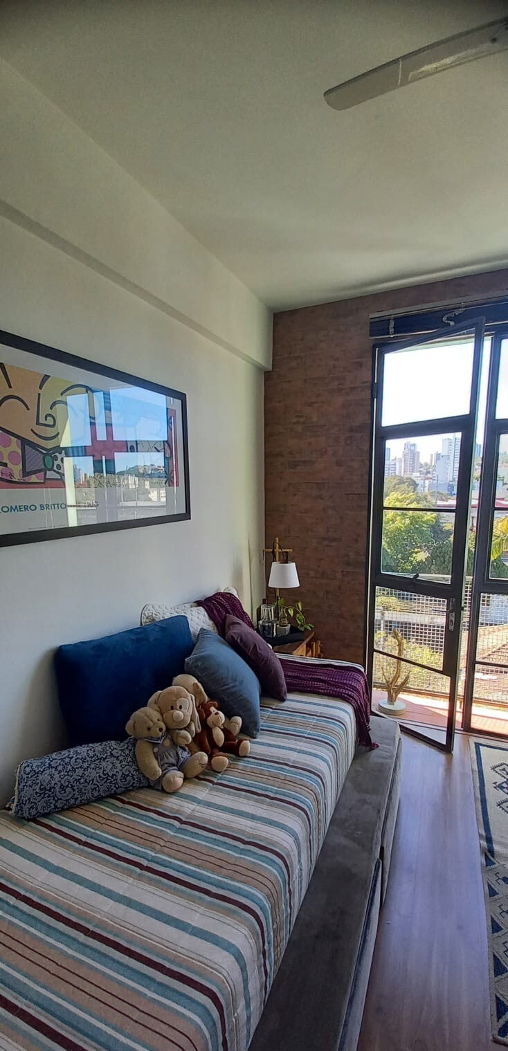 A cozy sleeping space features a striped bed adorned with a mix of pillows and two plush teddy bears. Natural light enters through the large glass door that opens to a view of the city and greenery, while a decorative wall art piece enhances the ambiance.