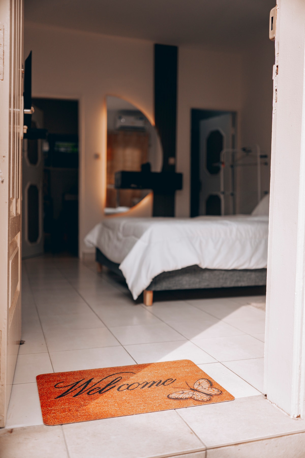 A welcome mat is placed at the entrance of a cozy room. The spacious bed is visible, dressed in crisp white linens. Soft lighting creates a warm ambiance, and the room features neutral-coloured walls with contemporary decor. The entrance is tiled, leading into a tidy living space.