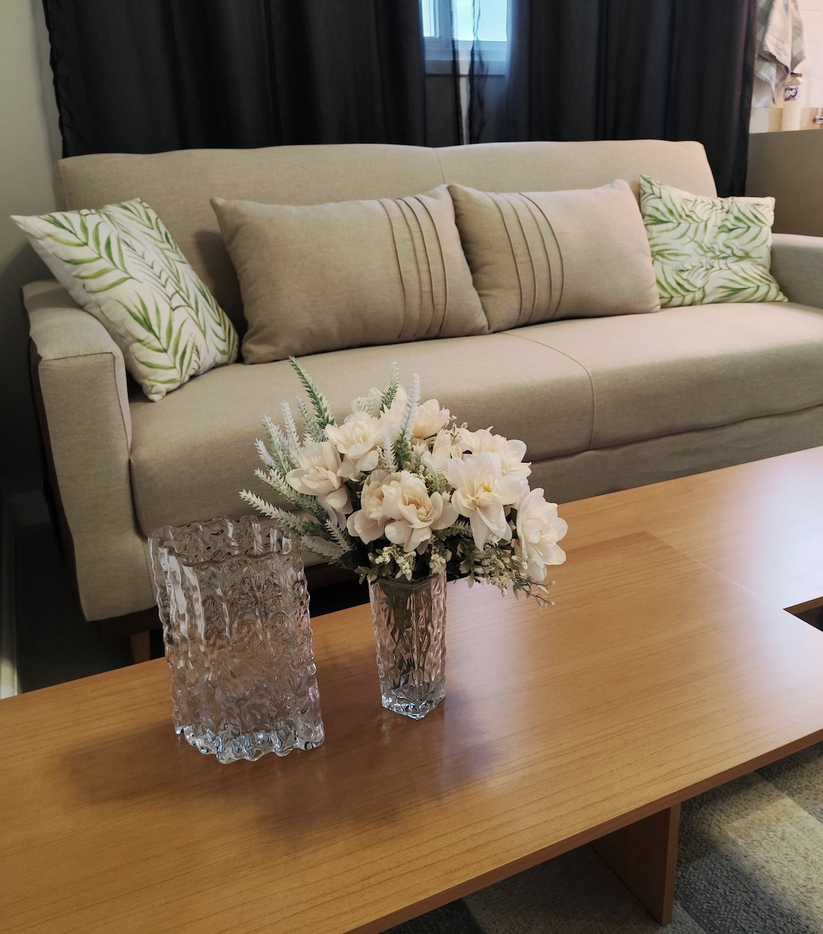 A light-toned sofa features decorative cushions with a botanical pattern, positioned against dark curtains. A wooden coffee table holds a textured glass vase filled with delicate white flowers and a crystal-like container, enhancing the room’s inviting design.