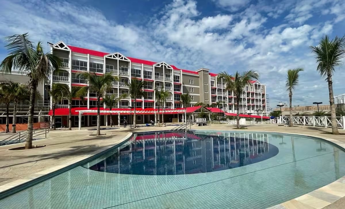 A contemporary building with multiple balconies is featured, accommodating palm trees in the foreground. A large swimming pool reflects the sky, promoting relaxation. The red accents of the structure contrast against the soft blue of the water, and a spacious deck surrounds the pool area.