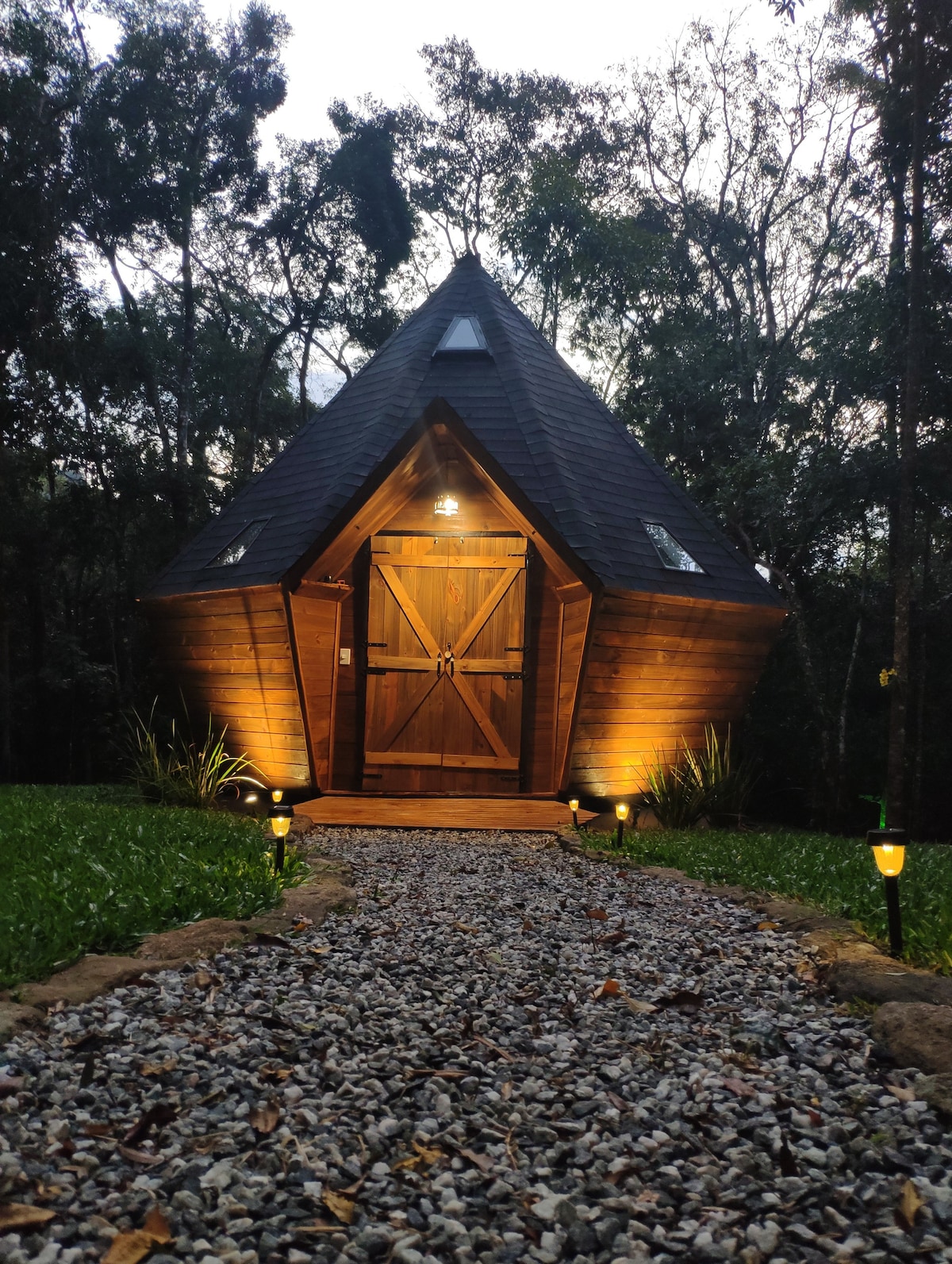 A diamond-shaped wooden cabin stands amidst lush greenery, illuminated softly by exterior lights. A gravel path leads to the welcoming front door, framed by well-maintained grass and subtle lighting along the walkway, creating a serene entrance to the natural surroundings.