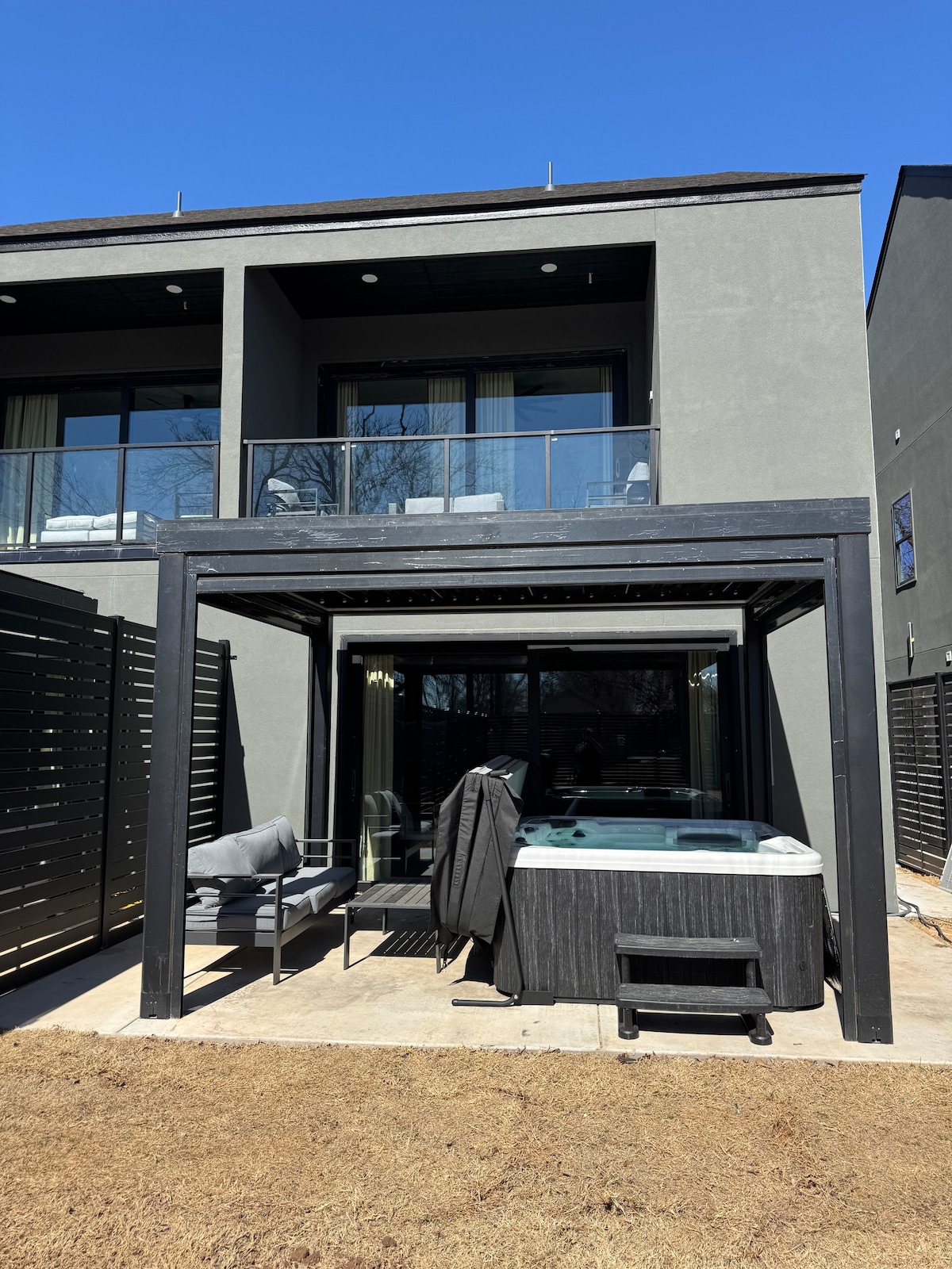 A modern outdoor space features a hot tub under a black pergola, accompanied by gray seating. Large glass doors lead into the home, allowing visibility of the interior. The area is illuminated by natural sunlight and surrounded by neatly arranged fencing.