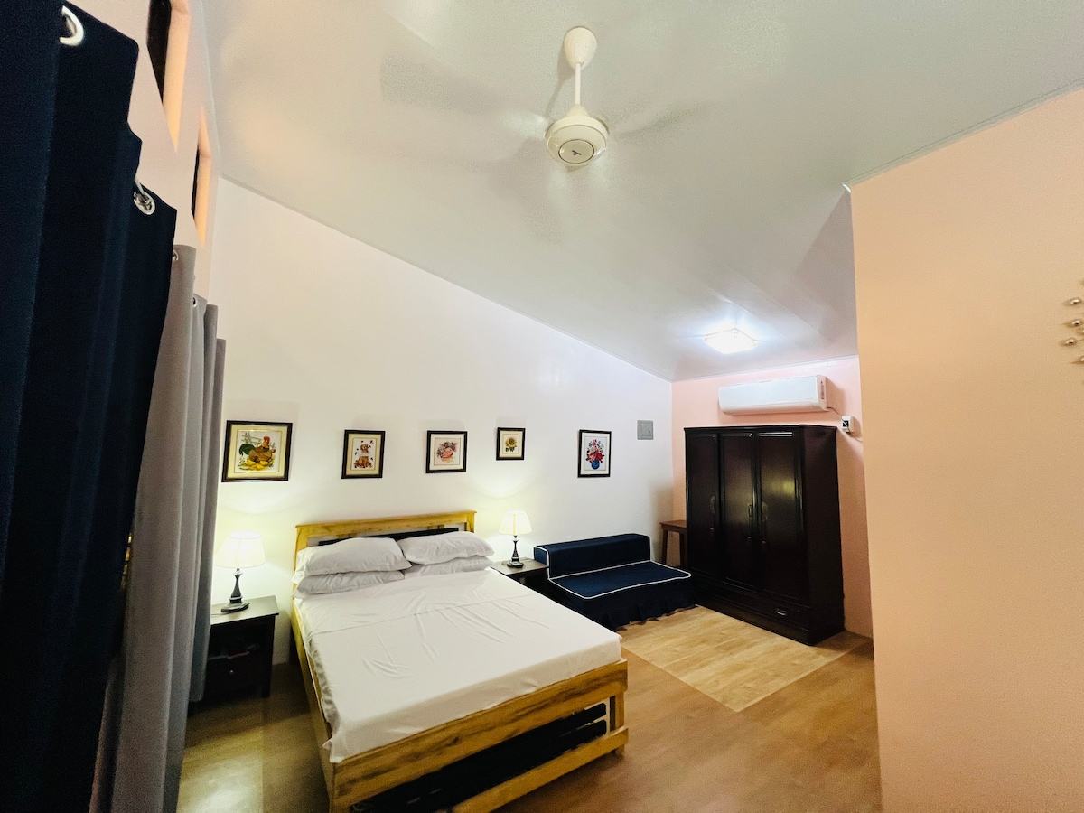 The Family Room displays a comfortable sleeping area featuring a spacious bed with neutral bedding. A sofa bed sits against one wall, complemented by a wooden cabinet. Framed artwork adorns the walls, and soft lighting from bedside lamps creates a cozy atmosphere.