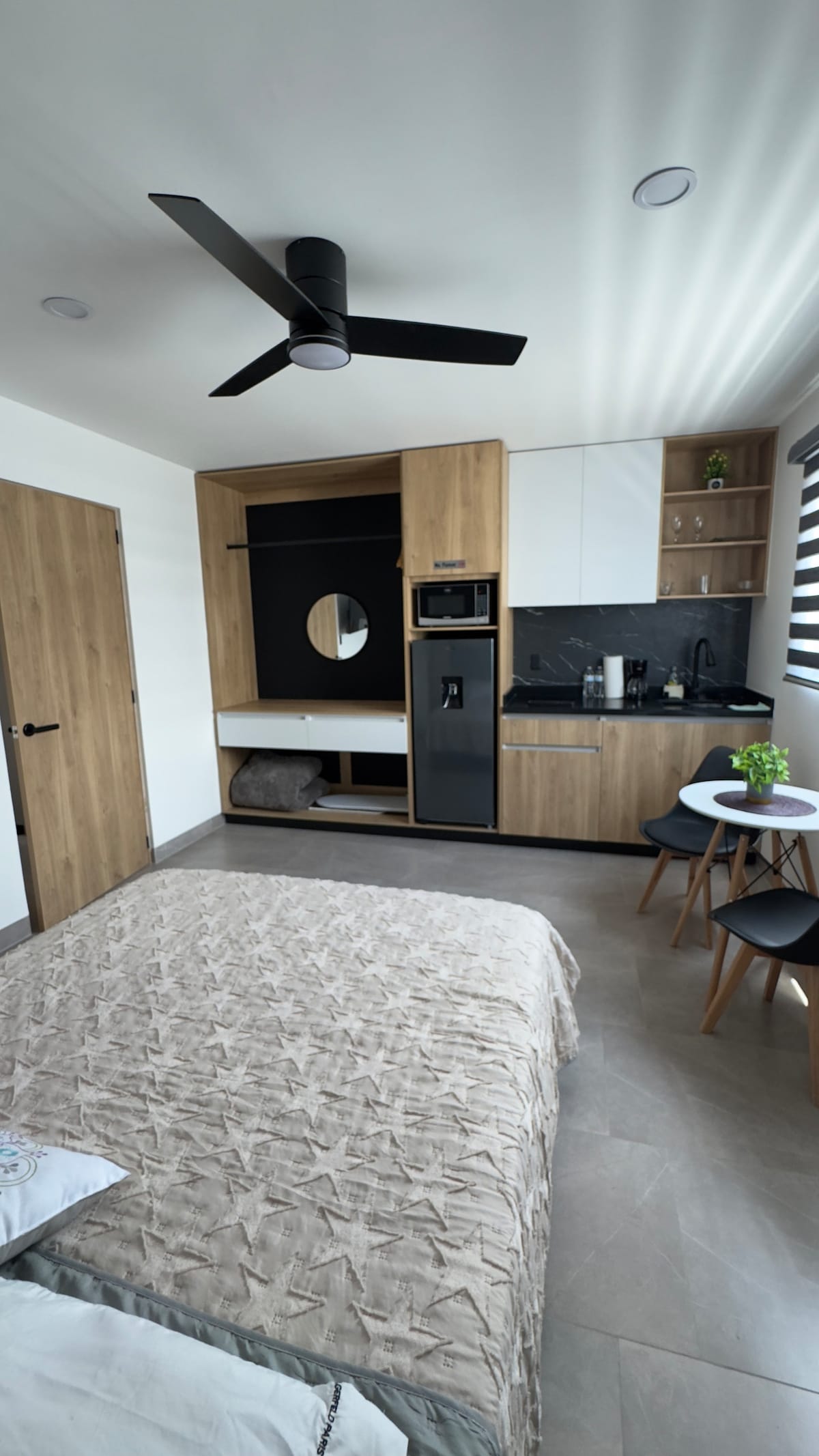 A modern loft features a spacious bed adorned with a textured coverlet. Built-in cabinetry and an integrated kitchen area are visible, alongside a round dining table with two chairs. Natural light filters through stylish window treatments, and a ceiling fan is present for comfort.