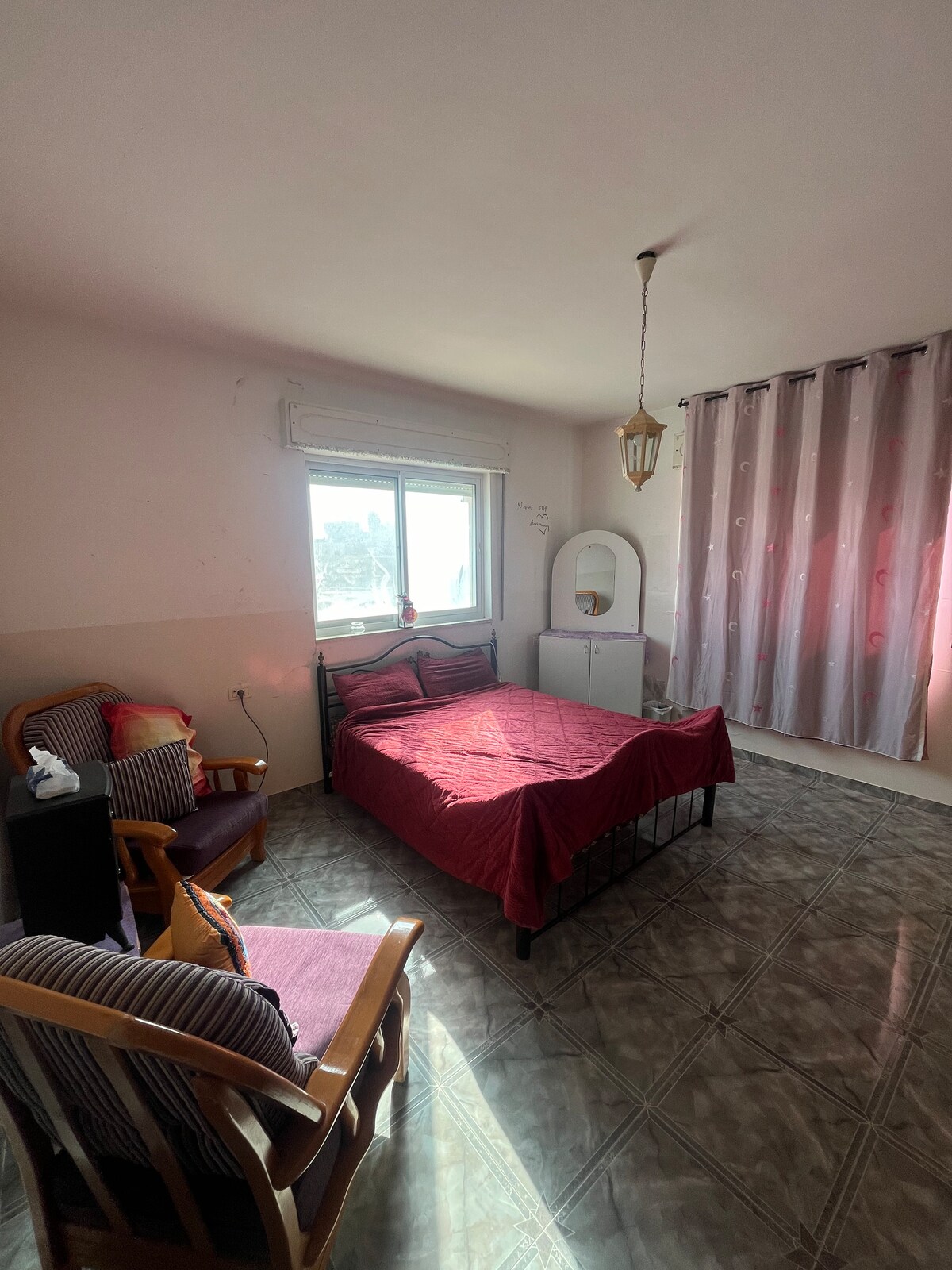 A bright bedroom features a spacious bed dressed in red linens, complemented by a small nightstand. Two armchairs are positioned nearby, creating a seating area. A window allows natural light to fill the room, while sheer curtains gently diffuse the light. A mirror and storage unit are present.