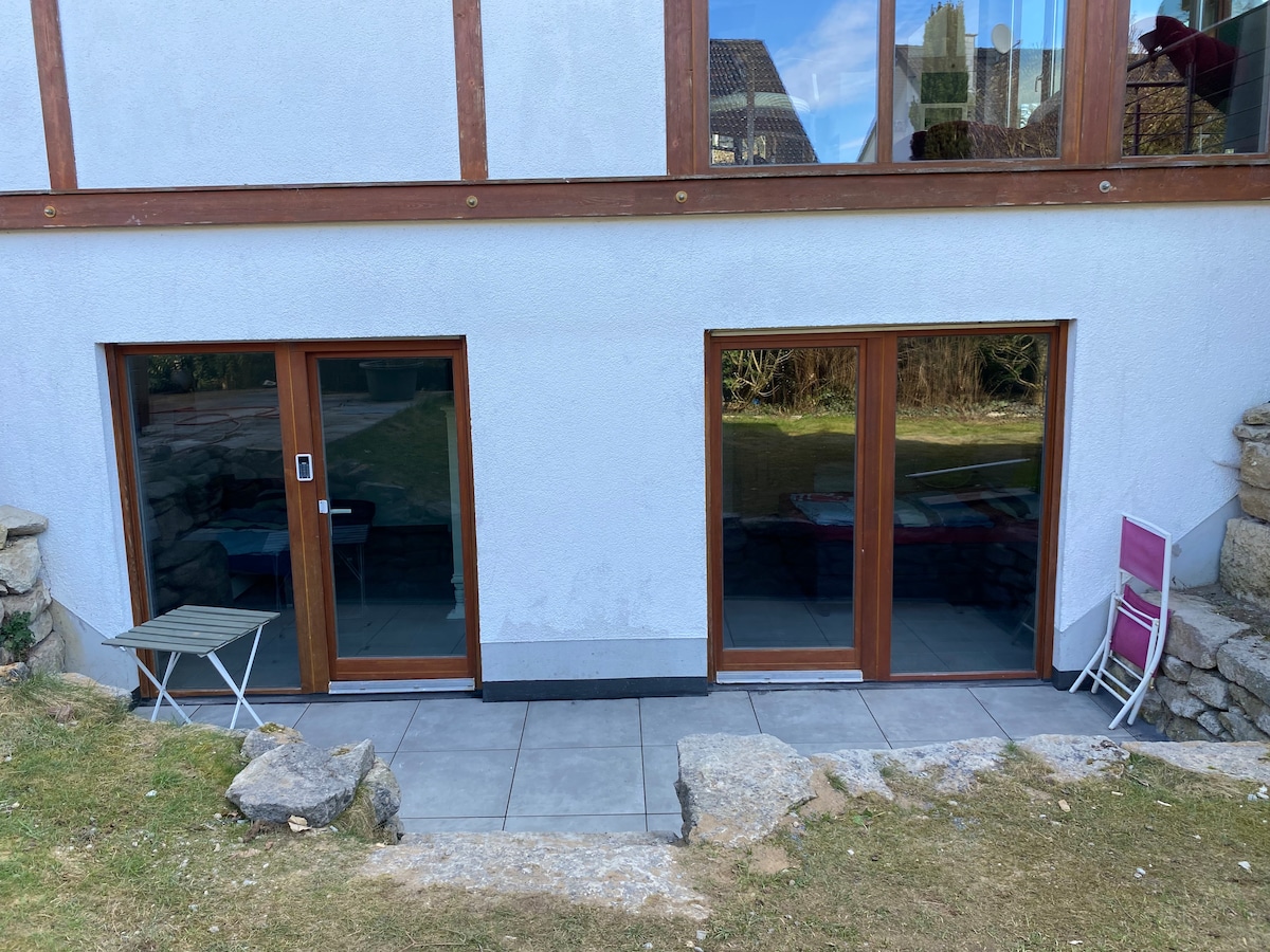 Two sets of glass doors are visible at the lower level of the building, leading to a covered patio area. The modern design includes a small table and a chair set beside the doors, with a well-maintained garden space extending beyond.