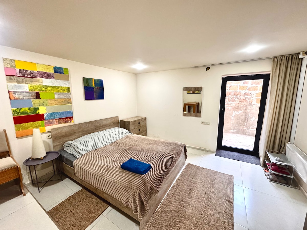 A spacious studio with a double bed is illuminated by overhead lights. A variety of colorful artwork decorates the white wall. A wooden dresser is placed beside the bed, while a mirror reflects the room. Large doors lead to an outdoor area with natural light flooding in.