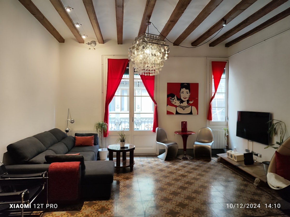 The modern living area features a dark sectional sofa with red accent pillows, complemented by a glass coffee table. A large chandelier hangs from the ceiling, while bright red curtains frame the large window. A colorful piece of wall art adds character beside the smart TV and two stylish chairs.