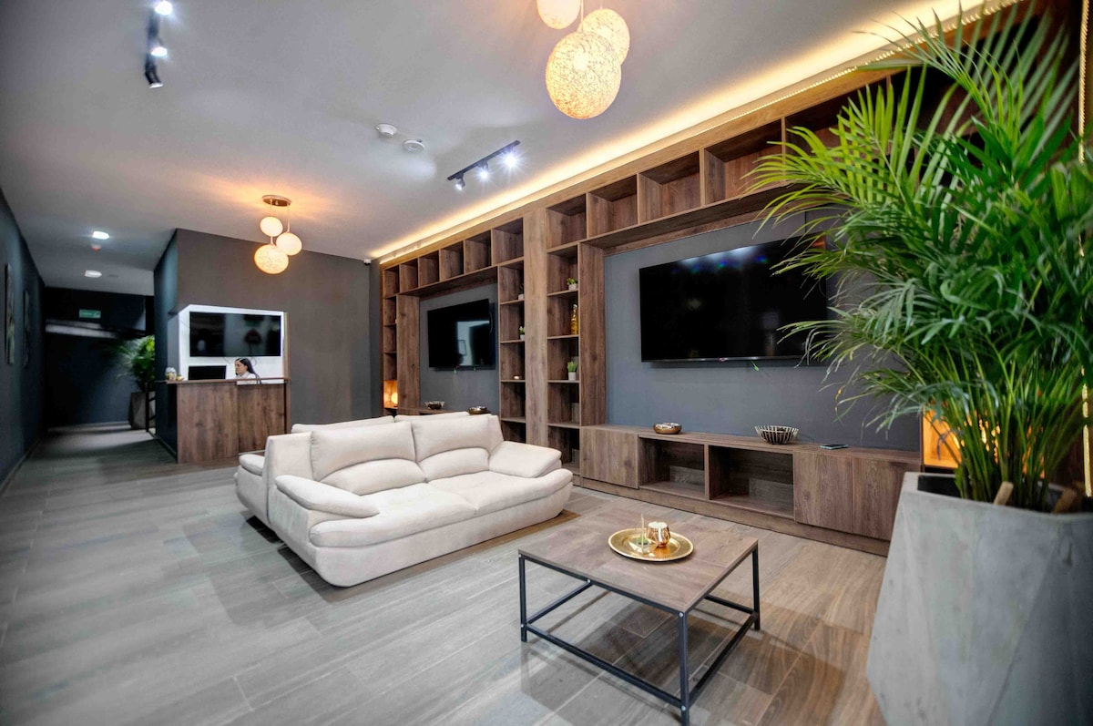 A modern lounge area is presented with soft seating arranged around a low coffee table. Wood shelving lines the walls, adorned with decorative items. Two flat-screen televisions are mounted, and a potted plant adds a touch of greenery.