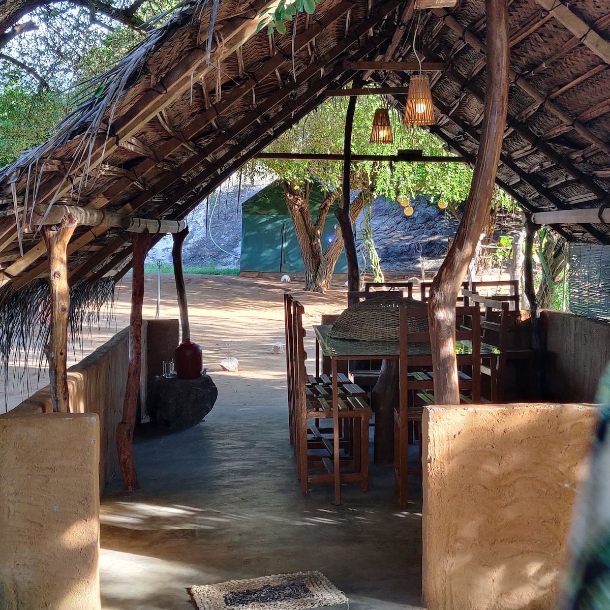 An open-air dining area features a thatched roof supported by wooden beams. A solid wooden table with several matching chairs occupies the space, complemented by soft lighting fixtures. Natural light filters through, illuminating the rustic walls and the surrounding greenery.