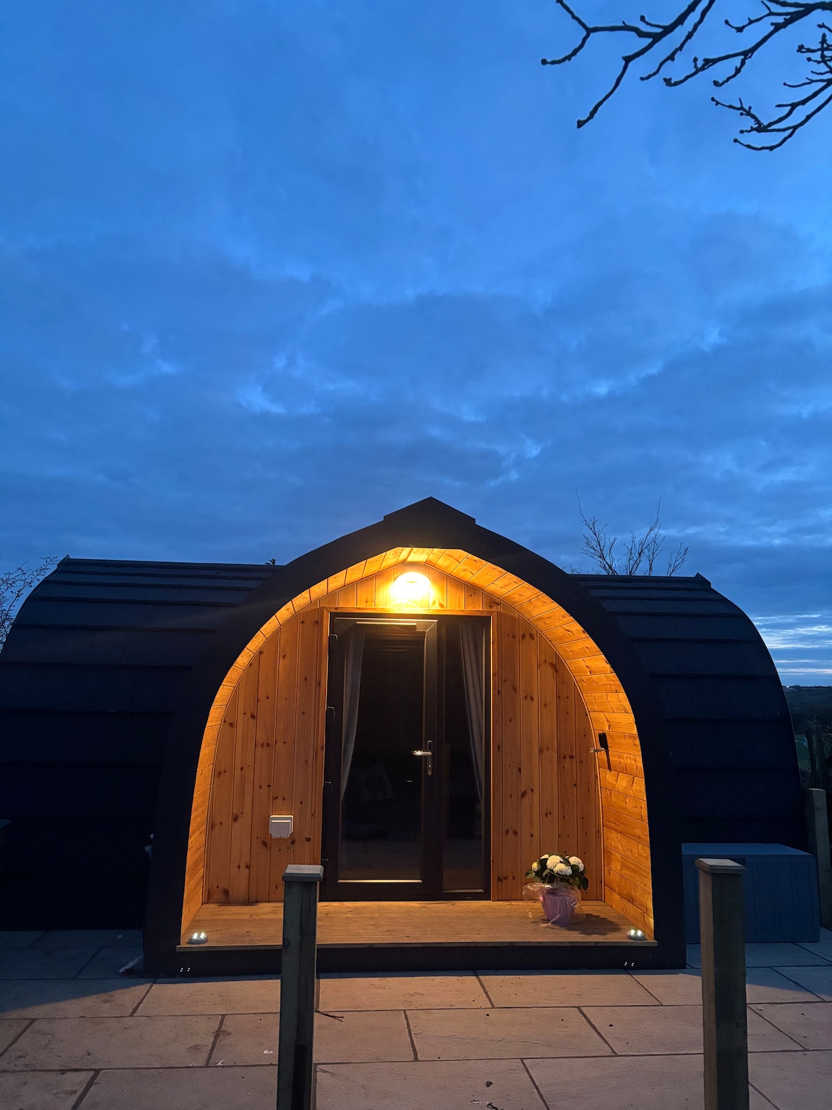 A charming glamping pod with a distinctive curved shape stands illuminated at dusk. Warm light radiates from the entrance, highlighting the wooden façade. A small potted plant is positioned by the doorway, creating a welcoming entrance at the end of a stone pathway.
