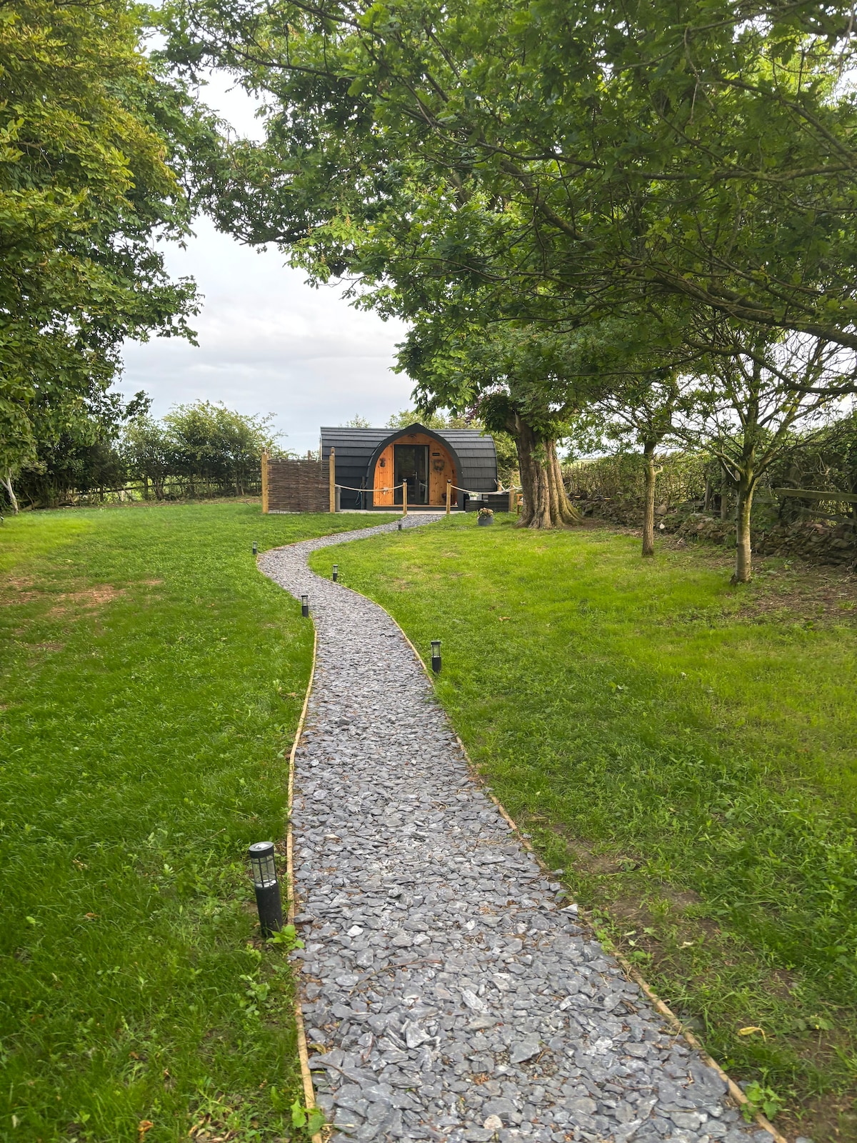 A gravel pathway leads gently through a lush green lawn towards 'The Highlander' pod, framed by trees and bushes. Soft lighting fixtures line the pathway, guiding guests to the entrance of the cabin, nestled in a serene natural setting.