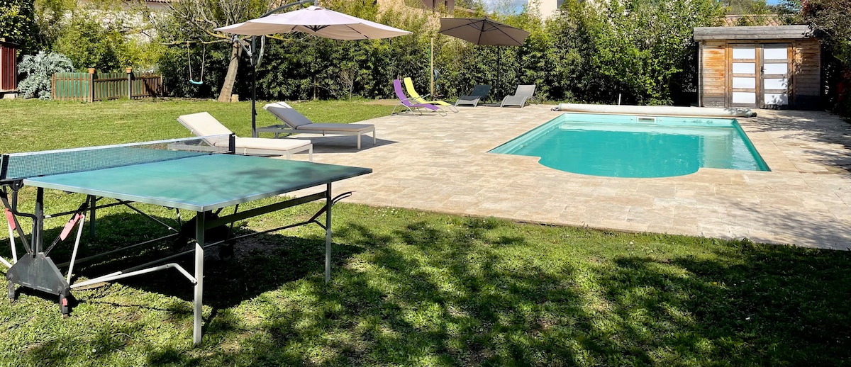 An outdoor area features a swimming pool surrounded by a travertine deck, lounge chairs, and umbrellas. A table tennis setup is positioned on the lawn, enhancing the recreational opportunities within the spacious garden, which is thoughtfully landscaped for relaxation.