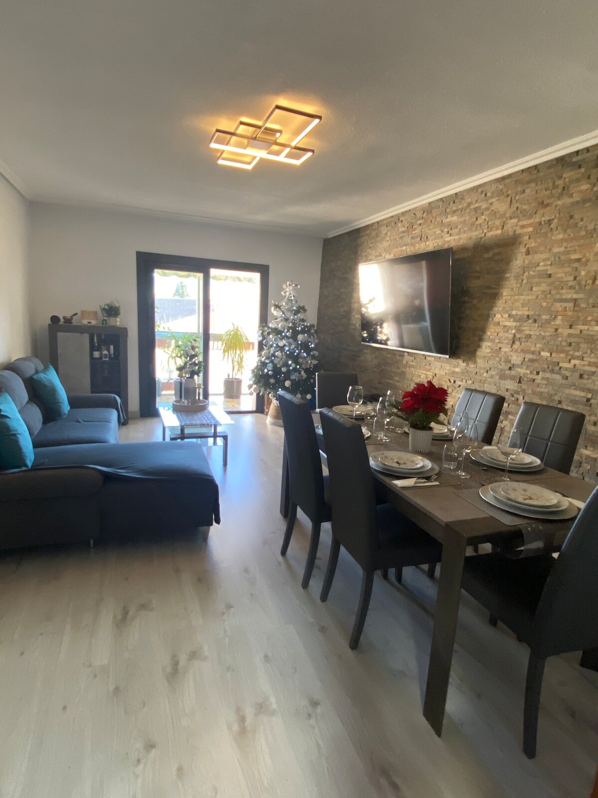A modern living space features a comfortable gray sofa adorned with teal pillows. A wooden dining table is set for six, accompanied by dark chairs. Natural light enters through glass doors that open to a balcony, while a Christmas tree adds a festive touch.