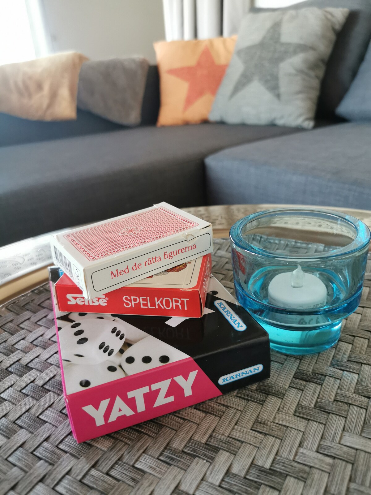 A cozy living area is depicted, featuring a gray sofa adorned with star-patterned cushions. A woven table holds a stack of board games, including a Yahtzy game and a deck of cards. A teal-colored candleholder contains a lit tea light, adding a subtle warmth to the space.