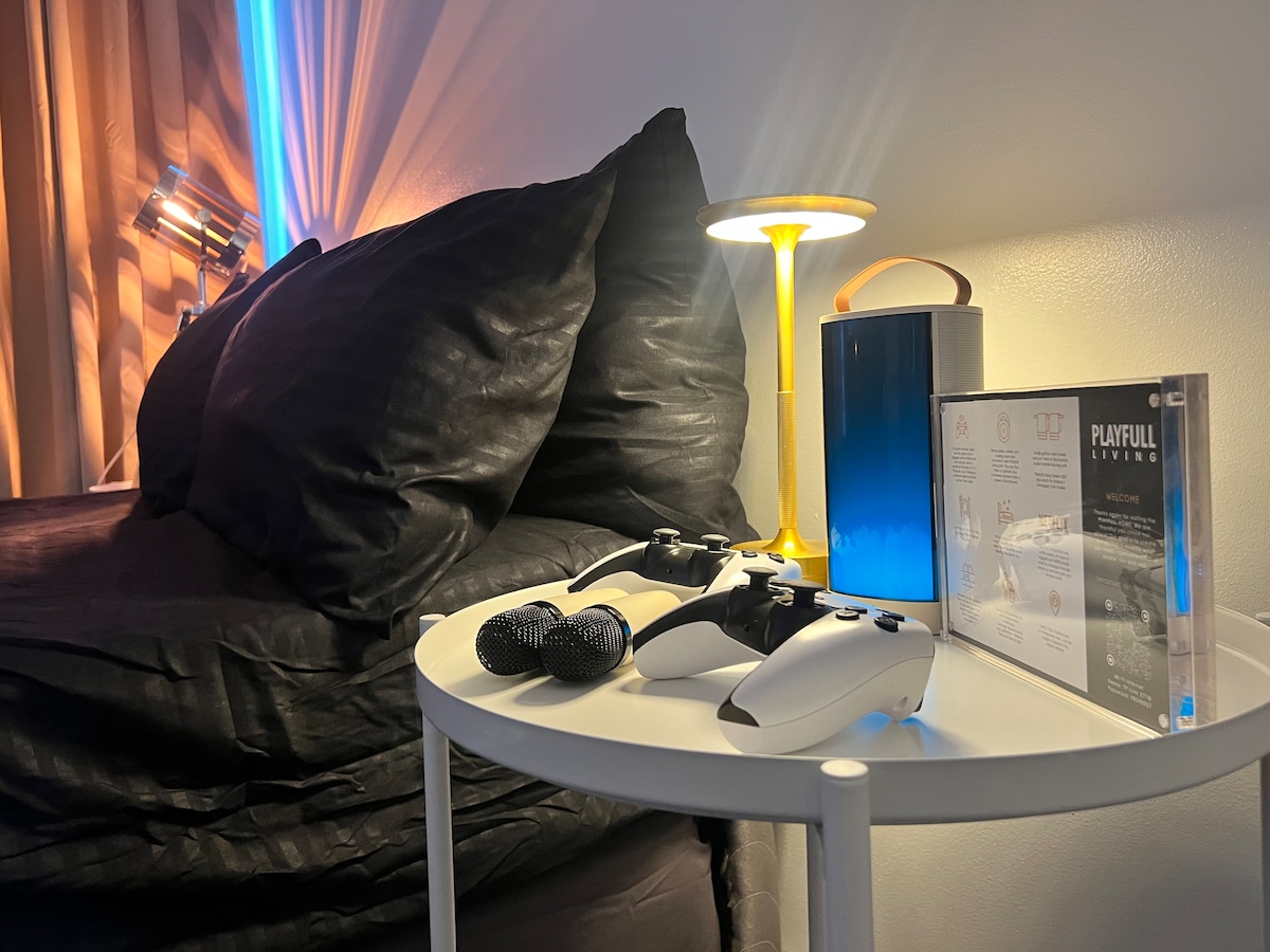 A bedside table is displayed with gaming controllers and a portable speaker. A colorful lamp provides soft lighting, while a compact console is positioned nearby. The adjacent bed is dressed in dark linens, creating a modern and relaxed ambiance.