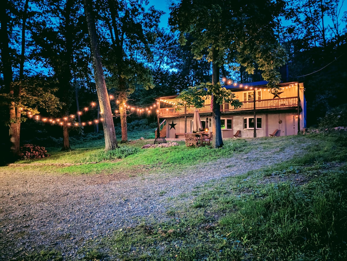An inviting retreat is set in a wooded area, illuminated by string lights that enhance the evening ambiance. The dwelling features a deck with chairs, and a gravel area is surrounded by greenery, creating a serene outdoor setting for relaxation.