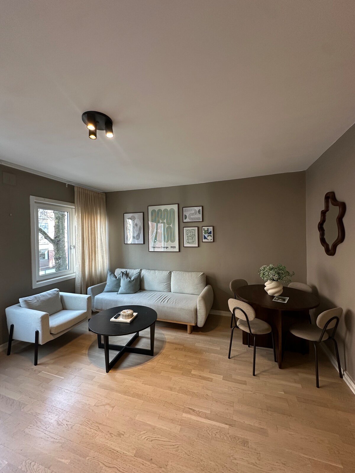 The living area features a light-colored sofa paired with a compact armchair. A round table with four chairs occupies one corner, while wall art and a decorative mirror add interest. Natural light filters through a window dressed with sheer curtains.