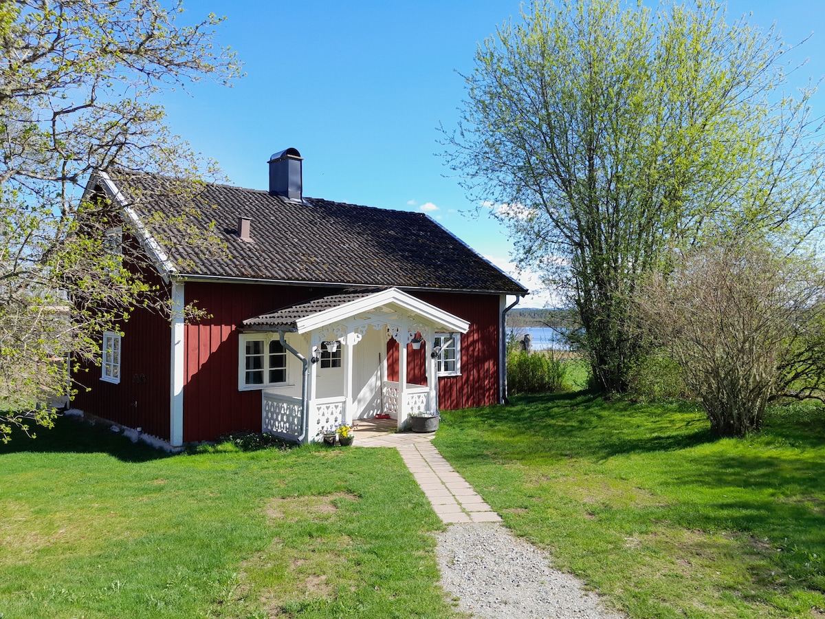 A charming red house with a gabled roof is situated in a grassy area, surrounded by trees. A stone pathway leads to a welcoming porch, while the serene lake is visible in the background, framed by budding greenery.