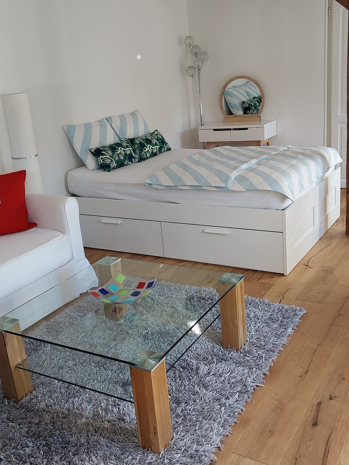 A bright sleeping area is presented, featuring a spacious bed adorned with striped bedding and decorative pillows. A glass coffee table is positioned on a soft rug, accompanied by a small lamp and a mirror on a bedside cabinet. A white couch adds seating to the space.