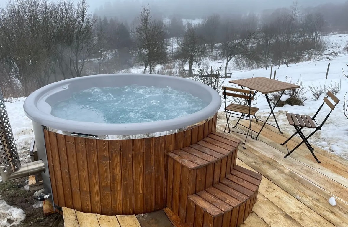 A wooden hot tub is surrounded by a snowy landscape, with mist rising from the warm water. Nearby, a small table with two chairs is positioned on wooden decking, providing a cozy outdoor seating area in a natural setting.