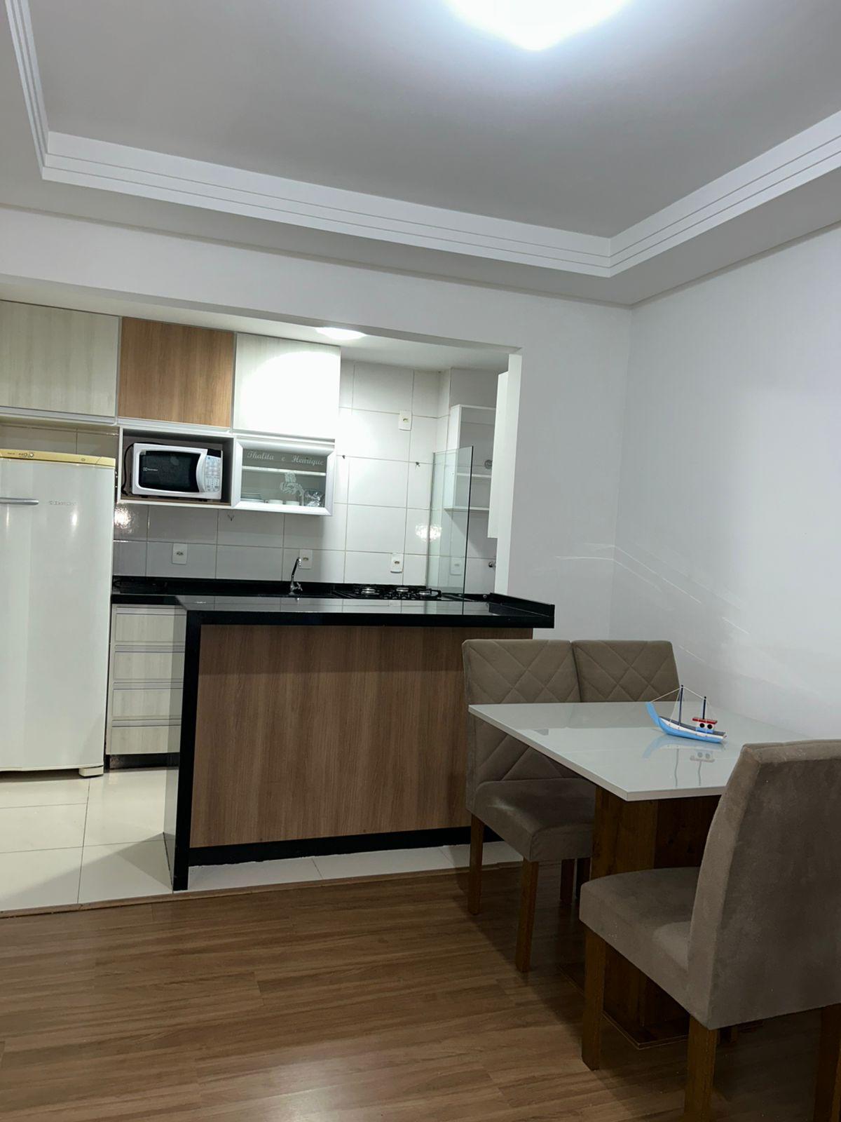 A contemporary kitchenette is visible, featuring light wood cabinetry and a black countertop. Modern appliances, including a microwave and refrigerator, are integrated into the design. A small dining area with a glass table and two upholstered chairs complements the space, creating a functional dining spot.