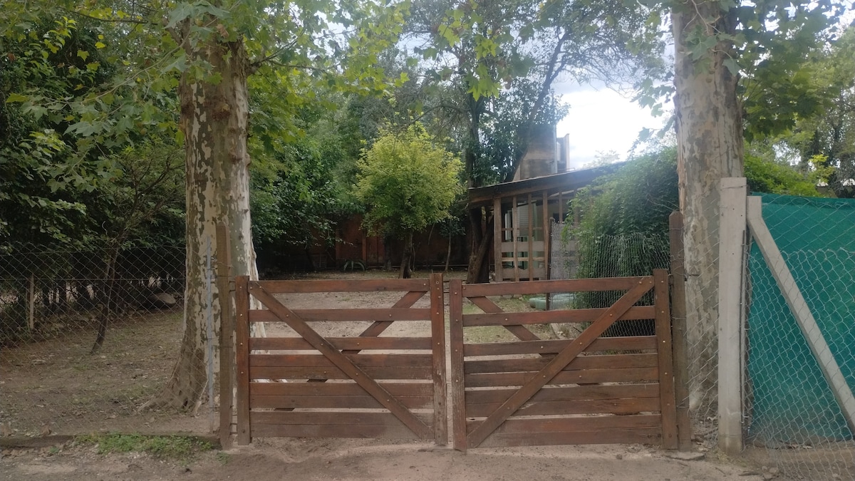 A wooden gate stands open, providing access to a property surrounded by lush greenery. Tall trees frame the entrance, while a cozy building is visible in the background. Natural light filters through the foliage, enhancing the tranquil setting.