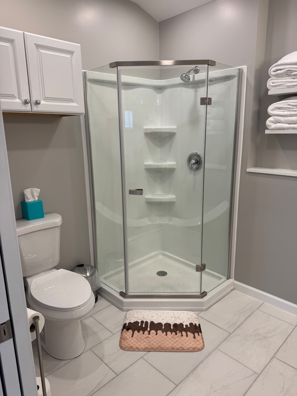 A compact bathroom is displayed, featuring a glass-enclosed shower with built-in shelves. A white toilet is positioned beside the shower, and a light-colored mat is placed on the floor. Shelving above holds neatly stacked towels, while a decorative tissue box adds a pop of color.