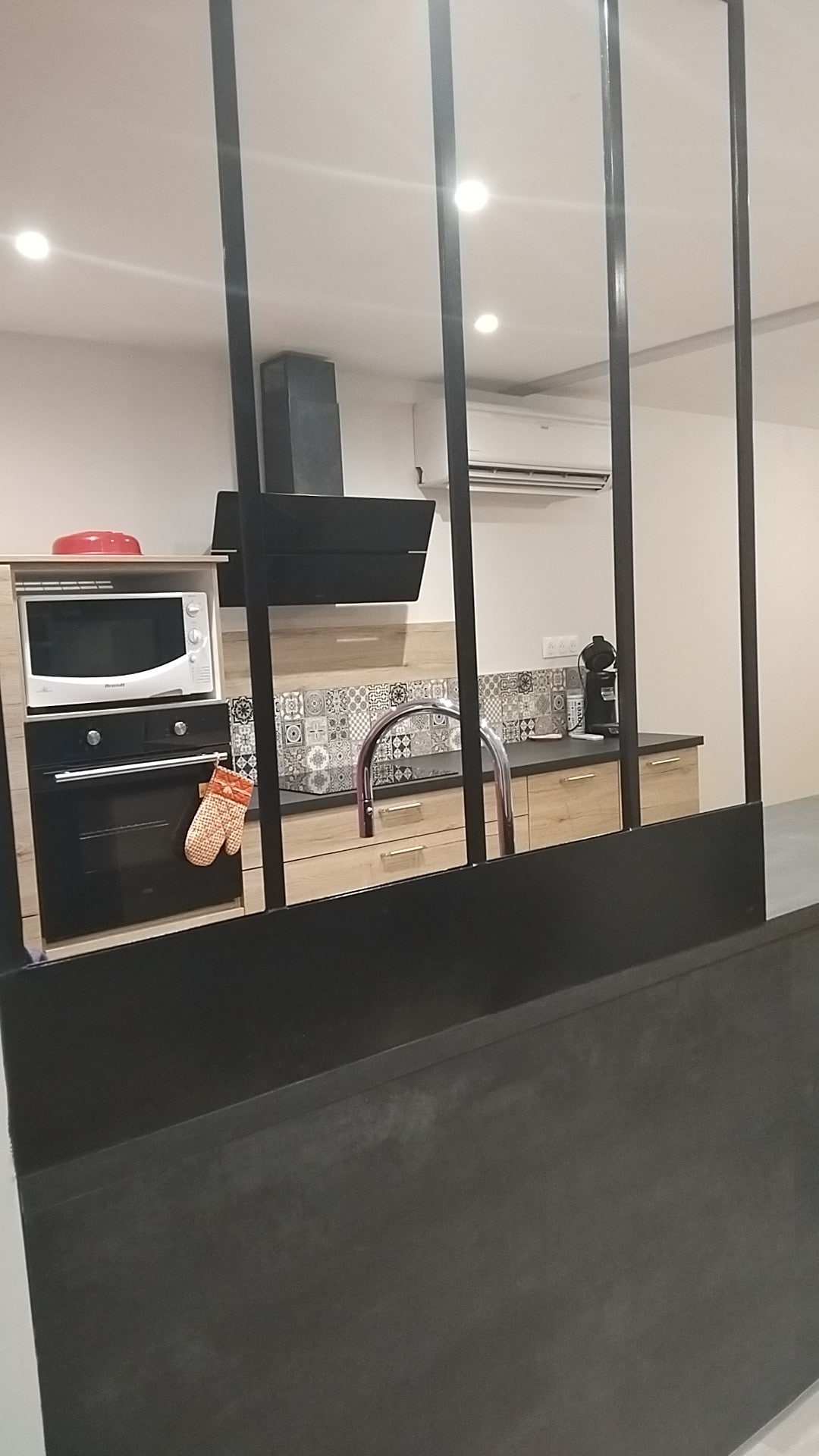 A modern kitchen area is visible through a sleek black partition. Features include an oven, microwave, and stovetop, with a patterned backsplash adding character. A sink and coffee maker are also present, along with a red oven mitt hanging from the counter.
