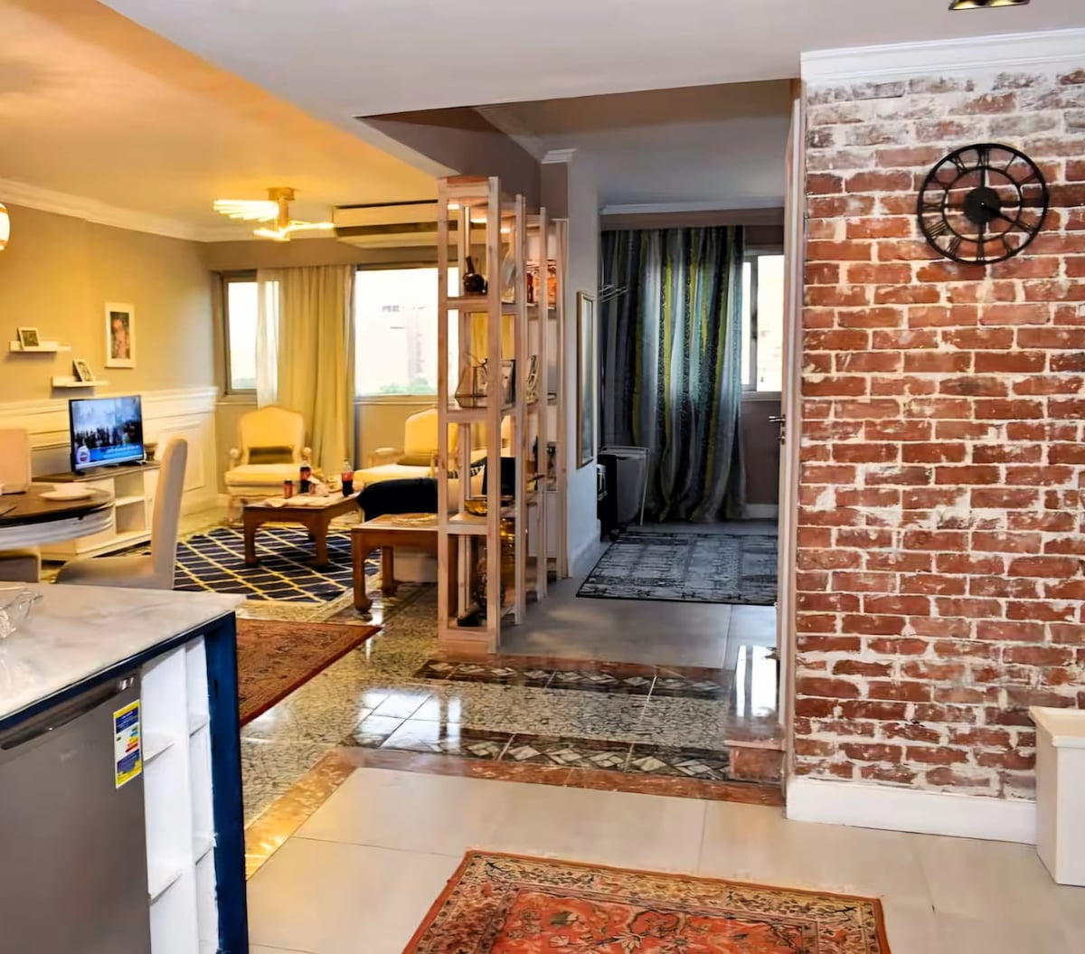 The open living area is visible, featuring a comfortable sofa and a dining table. Natural light is captured through large windows. A textured brick wall adds character, while decorative rugs enhance the space. The kitchen area appears well-equipped and seamlessly blends into the design.