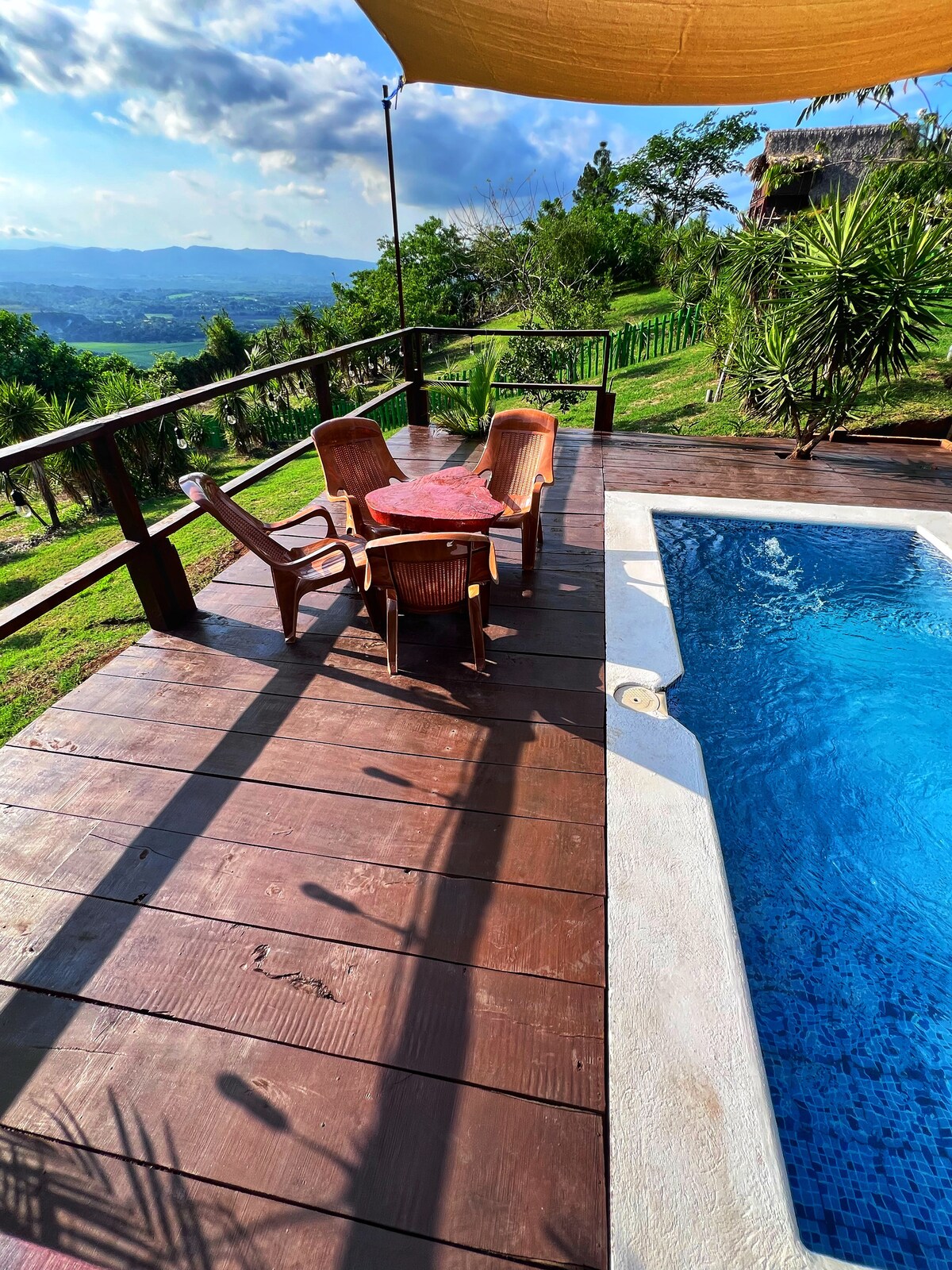 A wooden deck features a round table surrounded by four woven chairs, positioned beside a swimming pool. The clear water reflects the sky above, while lush greenery and expansive hills can be seen in the background, offering a serene outdoor setting.